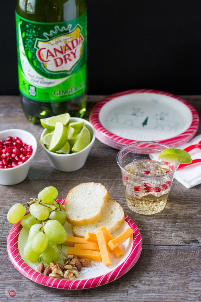 A wood table with paper plates, napkins, limes, pomegranate pieces, a drink and a bottle of canada dry ginger ale.