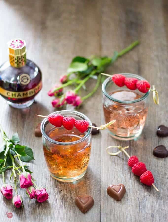 overhead of Chambord Raspberry liqueur, 2 raspberry love cocktails, chocolates and flowers on a wood surface