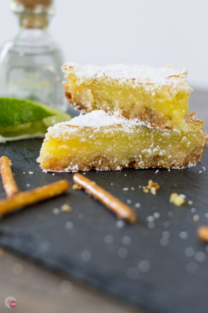 My favorite cocktail is now a dessert bar. Margarita Bars have a creamy and tart lime filling complete with a splash of tequila that sits atop a shortbread salty pretzel crust. A crispy top dusted with powdered sugar finishes off of this amazing dessert. Margarita Bars Recipe | Take Two Tapas | #MargaritaBars #Margarita #LemonBars #CincoDeMayo