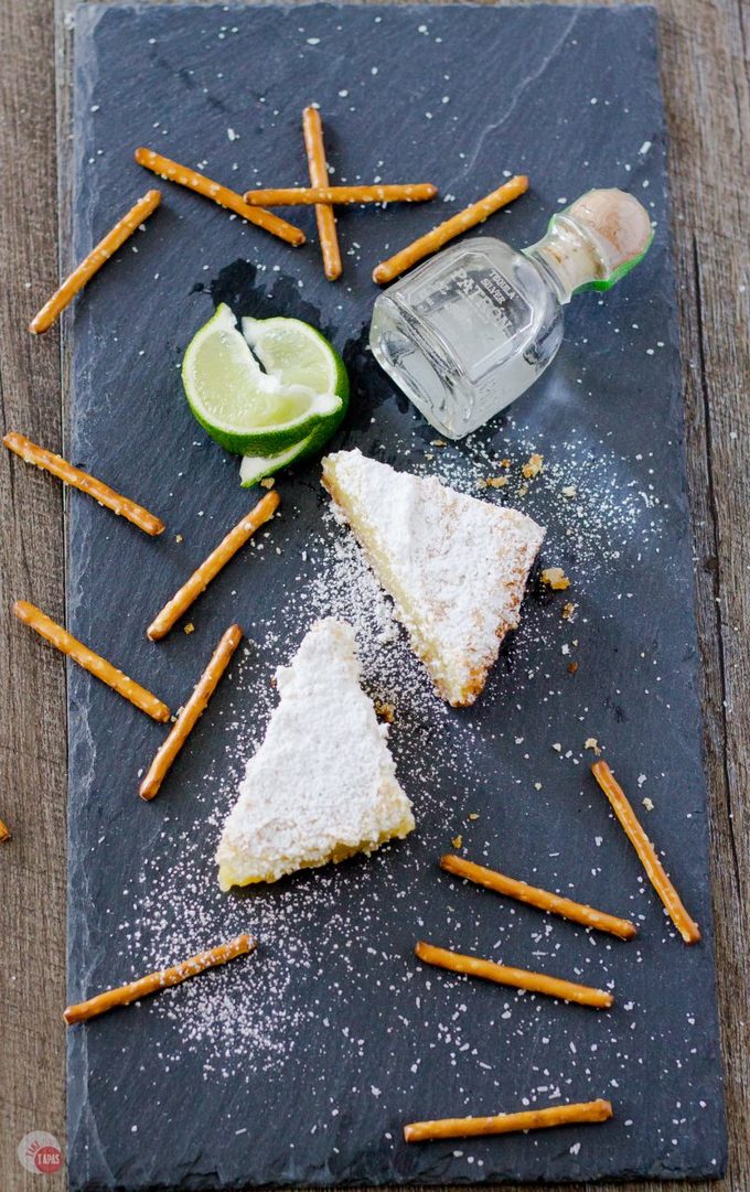 My favorite cocktail is now a dessert bar. Margarita Bars have a creamy and tart lime filling complete with a splash of tequila that sits atop a shortbread salty pretzel crust. A crispy top dusted with powdered sugar finishes off of this amazing dessert. Margarita Bars Recipe | Take Two Tapas | #MargaritaBars #Margarita #LemonBars #CincoDeMayo