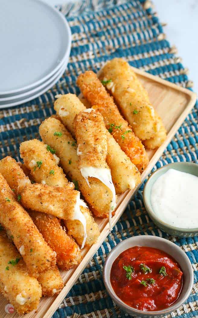 overhead picture of pile of cheese sticks with one broken in half and a small bowl of red sauce and a stack of plates