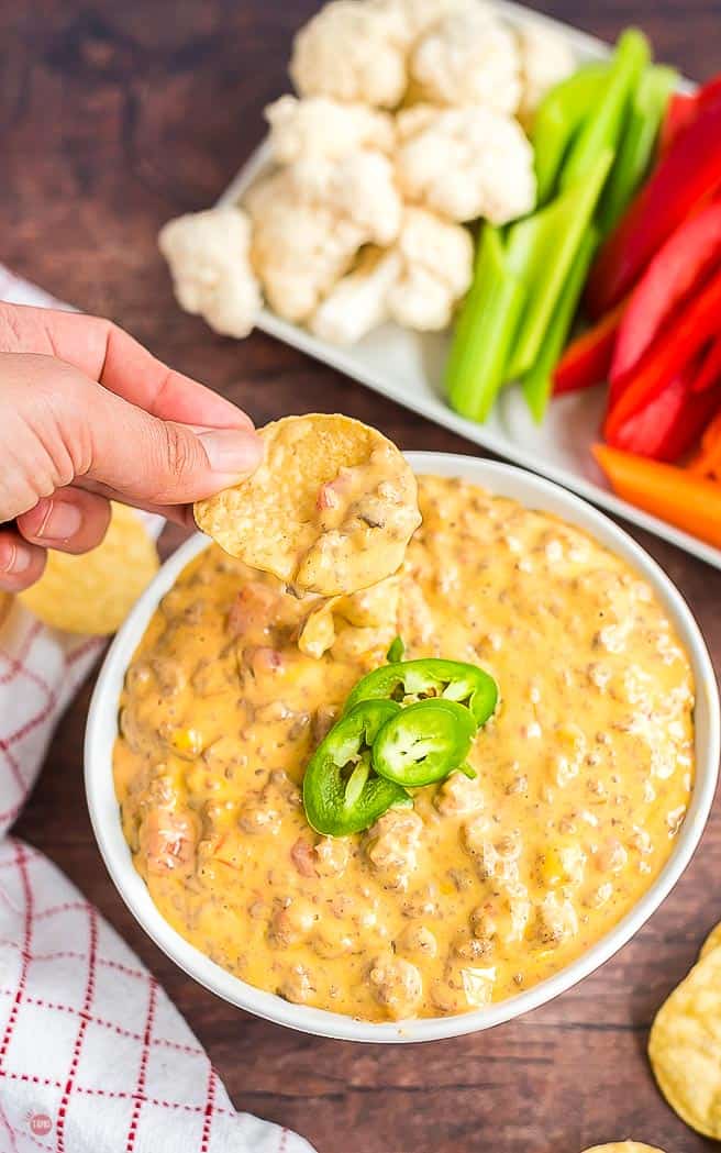 Chip schöpft etwas Käse-Dip in einer weißen Schüssel mit käsigem Rindfleisch-Dip mit Jalapenos mit einem Tablett mit Gemüse im Hintergrund