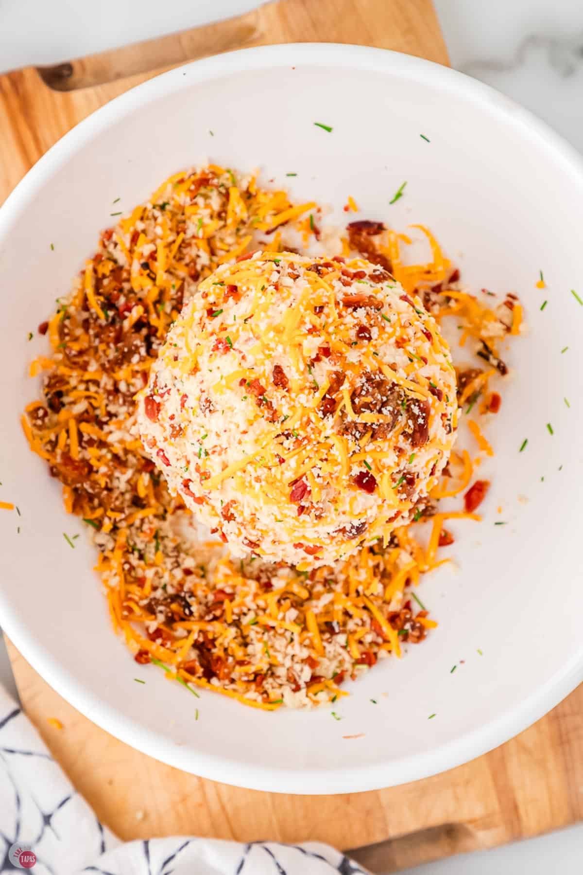 cheese ball being rolled in shredded cheese and bacon in a bowl